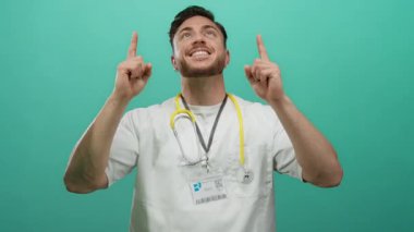 Steteskoplu genç beyaz erkek doktor yeşil bir arka plan duvarının önünde gülümsüyor ve yukarıya doğru işaret ediyor. Sağlık hizmetlerinde olumluluk ve iyimserliği yansıtıyor..