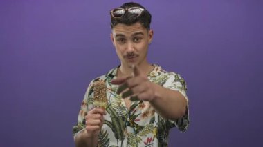 Tropikal tişörtlü genç İspanyol bir adam canlı mor bir arka planda dondurmanın tadını çıkarıyor, yüzünde bir gülümseme ve güneş gözlüğü ile şakacı hareketler yapıyor..