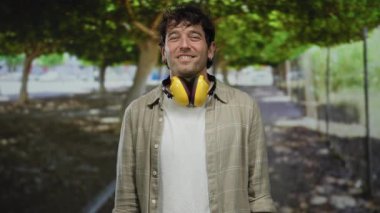 Yakışıklı görünümlü, kulaklık takan İspanyol bir adam, doğa tarafından çevrelenmiş dostça bir jest sergileyerek, ağaçlık bir sokakta elini uzatıyor..