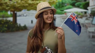 Dışarıdaki restoran terasında gülümseyen bir kadın elinde kameralı yeni Zelanda bayrağı ve açık havada neşeli bir seyahat anı sağlayan bir şapka takıyor..