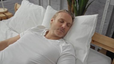 Orta yaşlı, beyaz bir adam, rahat bir yatak odasında uyanıyor, beyaz bir yatakta uzanıyor, evde sakin bir sabah rutini sergiliyor..