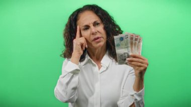 Orta yaşlı bir kadın yeşil bir geçmişe karşı İngiliz sterlini banknotları üzerinde kafa yoruyor, finansal karar verme ve tefekküre dalmayı somutlaştırıyor.