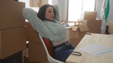 Yeni evindeki kutularla çevrili, masa başında yeni bir ev düzenini sergileyen, sandalyeye uzanmış bir kadın..