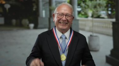 Takım elbiseli ve madalyalı bir adam sokakta, açık havada, gülümsüyor, halka açık bir ortamda bir numaralı ödülü gösteriyor..