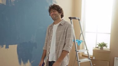 Genç adam, yeni oturma odasında merdiven ve kutularla duvarları boyuyor. Kapalı mekanların yenilenmesini öneriyor. Yüzünde şık bir gülümseme var.