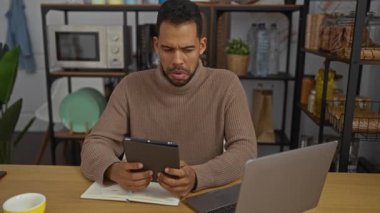 Genç adam rahat ofis ortamında tablet ve laptop kullanıyor. Arka planda raflar ve bitkiler var. Odaklanmış ve verimli bir iş yeri sahnesi oluşturuyor..