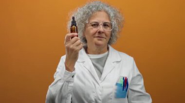 Laboratuvar önlüklü, gri saçlı orta yaşlı bir kadın damlatıcı şişeyi turuncu arka planda tutuyor, uzmanlık ve bilimi somutlaştırıyor..