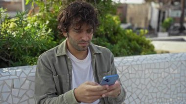 Şehir parkında akıllı telefonla konuşan genç bir adam, dışarıda yeşil alanlarla çevrili otururken şaşkınlığını dile getiriyor..