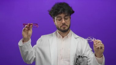 Sakallı genç adam laboratuvar önlüğündeki gözlükleri mor arka plandaki gözlüklerle karşılaştırıyor. Optikleri düşünceli bir ifadeyle ve modern tarzıyla inceliyor..