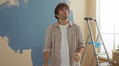 Evdeki duvarları boyayan genç adam, modern apartman ortamında arka planda doğal ışık altında sıradan bir gömlek giyen, düşünceli bir yüz ifadesiyle.