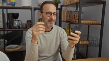 Raflar ve mutfak aletleri ile çevrili bir ofis ortamında akıllı telefonunu kullanarak bir video görüşmesine katılan yetişkin bir İspanyol..