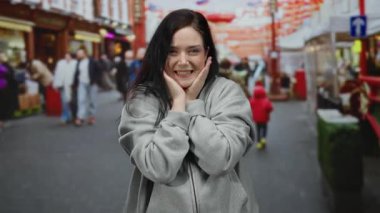 Kalabalık şehir ortamının ortasında gri kapüşonlu bir şey giyerken dışarıda el hareketi yapan bir kadın..