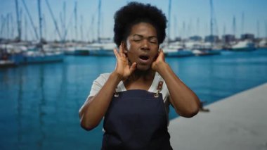 Önlüklü kadın, açık güneşli bir günde arka planda teknelerle sahil limanı yakınlarında açık hava çalışma ortamı sergileyerek el kol hareketi yapıyor..