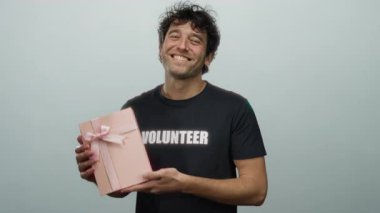 Gönüllü gömleği giyen gülümseyen adam gri arka plana karşı pembe hediye taşıyor. Cömertlik ve toplum ruhunu gösteriyor..