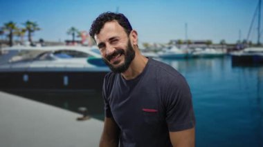 Sakallı İspanyol bir adam güneşli bir limanda sessiz kalmak için el kol hareketi yapıyor. Arka planda yatlar ve palmiye ağaçları var. Denizdeki sakin atmosferi vurguluyor..