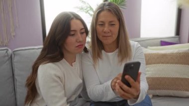 Oturma odasındaki kanepede kızına telefon gösteren bir kadın. Ev ortamında rahat bir aile bağı, teknoloji ve aşk anı yakalıyor..