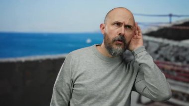 Sakallı kel bir adam, deniz kenarında bir arka planda dikkatlice dinliyor. Sahilde gezinirken düşünceli ve içsel bir tavır sergiliyor..