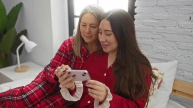 Anne ve kızı, rahat bir yatak odasında pijamalarıyla bir yatakta oturmuş sıcak bir iç mekanla sevgi, aile ve birliktelik hissini iletiyorlar..