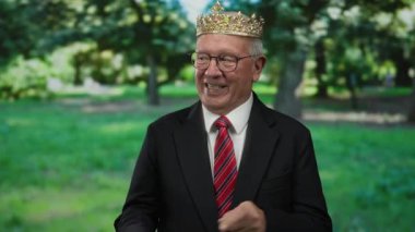 Takım elbiseli üst düzey bir adam taç takıyor ve açık hava parkında neşeyle işaret ediyor..