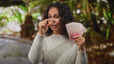 Tayvan banknotları tutan orta yaşlı bir kadın, yemyeşil bir park ortamında küçük bir el kol hareketi yapıyor, para ve finansmanı ön plana çıkarıyor..