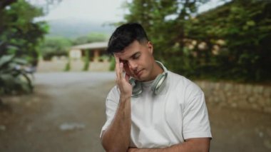 Kulaklıklı genç bir adam parkta dalgın dalgın duruyor. Doğal ve düşünceli ifadeler sergiliyor..