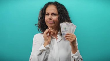 Orta yaşlı bir kadın elinde Kolombiya pezosu tutarken düşünceli bir şekilde mavi bir arkaplanda mali durumu ve parasal değeri yansıtıyor..