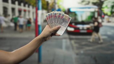 Açık hava otobüs durağında elinde parlak banknotlar, bulanık bir otobüs ve güneşli bir günde arka planda insanlar tutan bir adam..