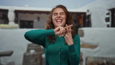 Sokak ortamında yeşil süveter giyen, şehir yaşamını ve canlı ifadeyi yansıtan beyaz arka planda gülümseyen hashtag jesti gösteren bir kadın..