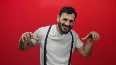 Sakallı, beyaz gömlek ve pantolon askısı giymiş, neşeli ve enerjik bir hava saçan, neşeli kırmızı bir arka planda poz veren İspanyol bir adam..