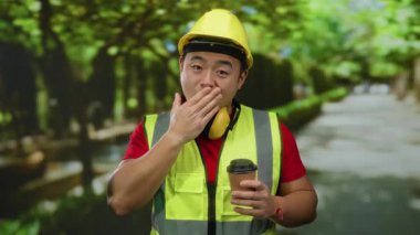 Yansıtıcı yelek ve sert şapka giyen genç bir adam elinde kahve tutarak açık hava parkında şaşırdı..