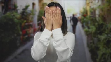 Genç Çinli kadın neşeli bir şekilde yüzünü gösteriyor, hareketli bir şehir ortamında kentsel bitki örtüsüyle çevrili canlı bir sokakta gülümsüyor, neşeli ve çekici bir açık hava sahnesi yaratıyor..