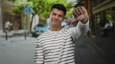 Çizgili gömlekli genç adam şehir caddesinde bulanık ağaçlar ve insanlarla başparmak kaldırıyor. Açık hava ortamını tasvip etmiyor veya hayal kırıklığına uğratmıyor..