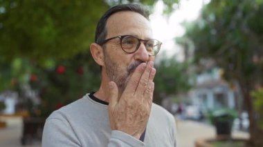 İspanyol bir adam, şehir parkında gülümseyip el sallıyor, orta yaşlı cazibesini, gözlüğünü ve sakalını açık hava ortamının ortasında sergiliyor..