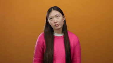 Pembe kazaklı, uzun saçlı bir kadın turuncu arka planda duruyor ve izole duvarın üzerinde derin derin bir ifade var..