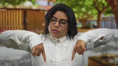Genç bir kadın park ortamını işaret ediyor, gözlüklü ve beyaz gömlekli, ağaçlarla çevrili, kendinden emin ve açık hava havasını gösteriyor..
