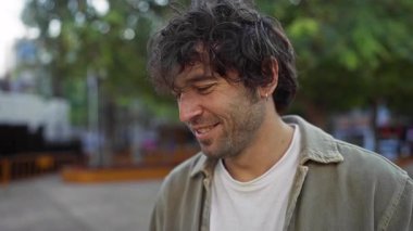 Arka planda ağaçlarla dolu bir şehir parkında gülümseyen genç adam canlı ve kaygısız bir şehir atmosferini yakalıyor.
