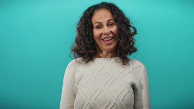 Kıvırcık saçlı, mavi arka plan duvarında gülümseyen, süveter giyen, stüdyo ortamında duygularını ifade eden olgun İspanyol bir kadın..