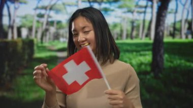 Açık hava parkında elinde İsviçre bayrağı tutarken gülümseyen bir kadın. Etrafı gür ağaçlarla çevrili. Genç Asyalı yetişkinlerin mutluluğunu ve kültürel gururunu temsil ediyor..