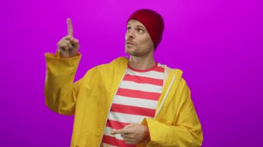 Sarı yağmurluklu ve pembe arka planda kırmızı şapkalı genç adam, karşıt ifadeler ve jestler sergiliyor, canlı moda ve İspanyol mirasını ön plana çıkarıyor..