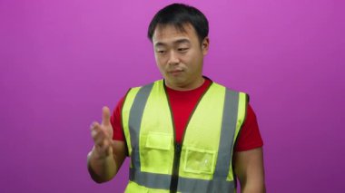 Emniyet yeleği giyen genç adam avuçlarını pembe arka plana dayamış gülümsüyor.