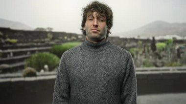 Gri kazaklı, kahverengi saçlı genç bir adam bir şehir caddesinde, bir parmak tabancası jesti yapıyor, can sıkıntısı ya da hüsran ifadesi sergiliyor..