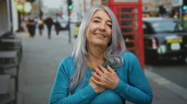 Meşgul bir Londra caddesinde, klasik bir kırmızı telefon kulübesinin önünde, elleri göğsüne yapışık bir şekilde duygularını ifade eden gri saçlı son sınıf öğrencisi bir kadın..