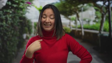 Parkta gülümseyen kırmızı kazaklı genç Çinli kadın kendini ağaçlar ve çalılarla göstererek açık havada hareketli bir ortam yaratıyor..