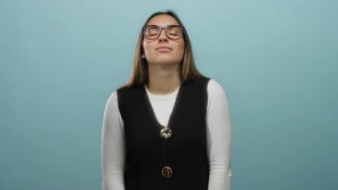 Gözlüklü ve yelekli kadın, izole edilmiş mavi arka plan duvarına karşı derin derin nefes alıyor, sakin ve odaklanmış, dingin bir atmosferde farkındalık ve rahatlamayı somutlaştırıyor..