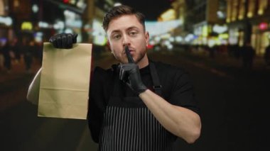 Sakallı, önlüklü ve eldivenli genç bir adam şehir merkezindeki açık hava ortamında sessiz bir jest yapıyor..