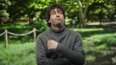 Güneşli bir günde yemyeşil bir parkta poz veren, parlak ağaçlar ve yaprakların arasından süzülen güneş ışığıyla çevrili gri bir kazak giyen düşünceli bir genç adam..