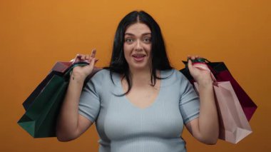 Parlak turuncu arka planda elinde alışveriş torbalarıyla gülümseyen bir kadın neşeli bir perakende terapisini temsil ediyor..