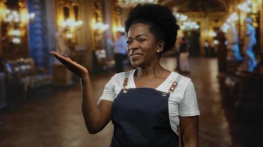 Zarif bir müze ortamında önlük giymiş, gülümseyen bir kadın. Arka planda sıcak ışıklar ve heykeller var..