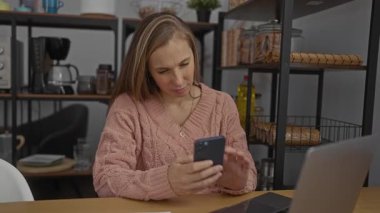 Kapalı bir ofiste dizüstü bilgisayarla çalışan bir kadın. Etrafı raflarla çevrili. Elinde cep telefonuyla örgü örülü bir kazak tutan bir kadın..