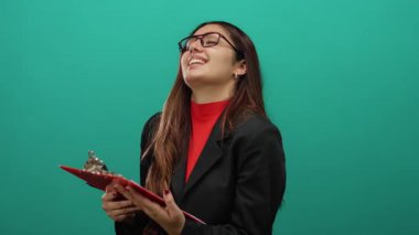 Canlı yeşil arka plana karşı elinde panoyla güvenle gülümseyen kadın, Latin kökenli ve iş ortamı için profesyonel kıyafetler sergiliyor.